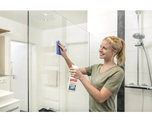 Femme nettoie une cabine de douche avec un nettoyant pour vitres et un chiffon dans une salle de bains.