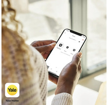 Eine Person hält ein Smartphone mit der Yale Home App zur Steuerung einer Alarmanlage in der Hand. Yale Home Logo.