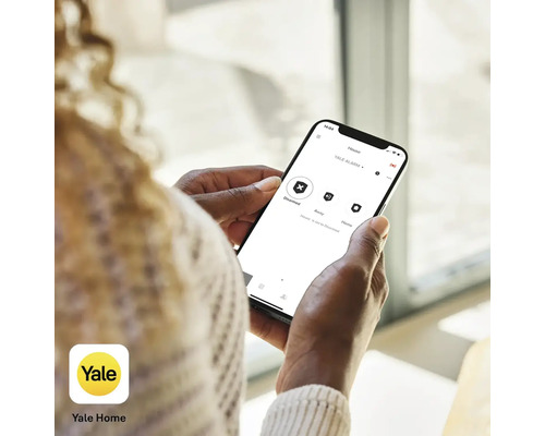 Eine Person hält ein Smartphone mit der Yale Home App zur Steuerung einer Alarmanlage in der Hand. Yale Home Logo.