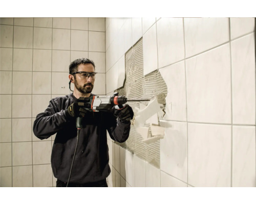 Un homme portant des lunettes de protection et des gants retire des carreaux d''un mur avec un marteau perforateur.