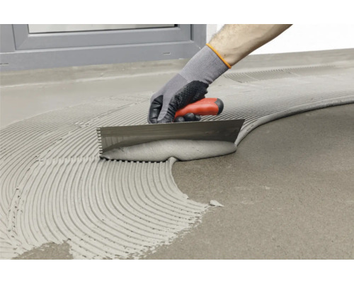 Handwerker verteilt Fliesenkleber mit einer Zahnspachtel auf einem Untergrund.
