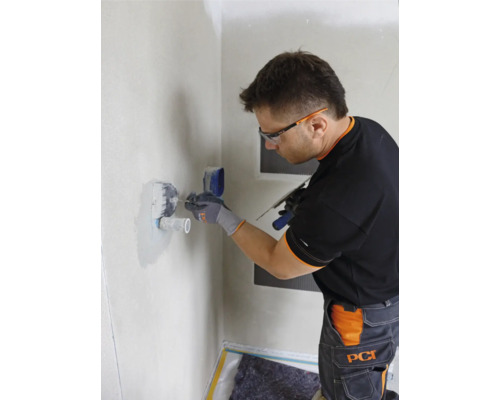 Ein Mann mit Schutzbrille arbeitet mit einer Spachtel an einer Wand im Badezimmer.