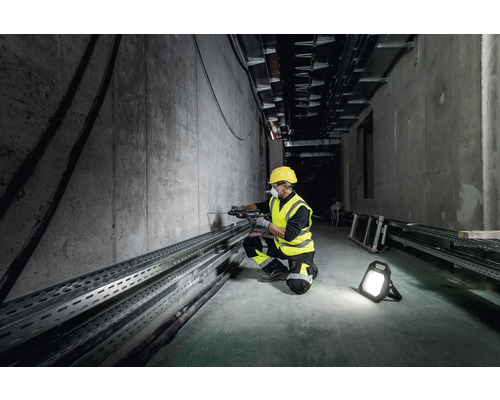 Un ouvrier portant un casque et un gilet de sécurité perce un mur en béton à l''aide d''une perceuse, éclairé par un projecteur de chantier.