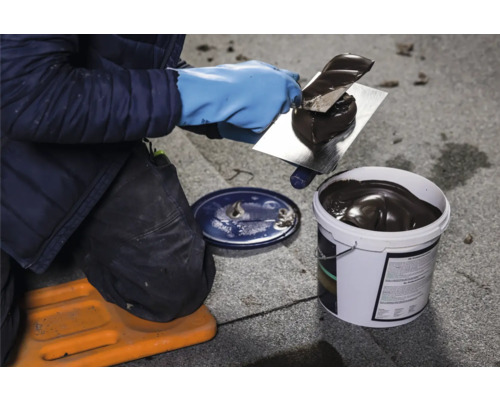 Eine Person trägt mit einer Kelle Dachkleber auf ein Dach auf, der Kleber stammt aus einem Eimer.