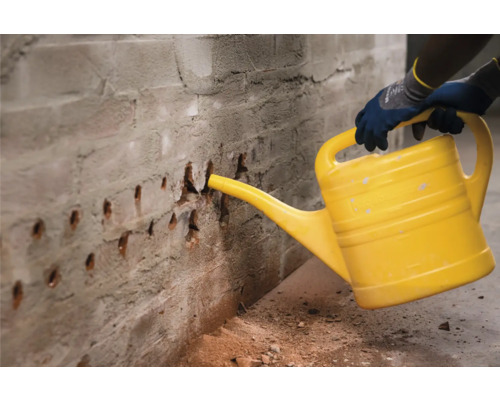 Eine Person befeuchtet Bohrlöcher in einer Ziegelwand mit einer gelben Gießkanne.