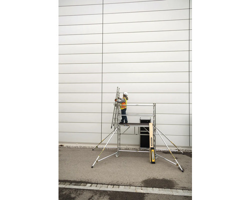 Eine Person mit Schutzhelm und Sicherheitsweste steht auf einem Rollgerüst und hält eine Leiter an einer Wand.