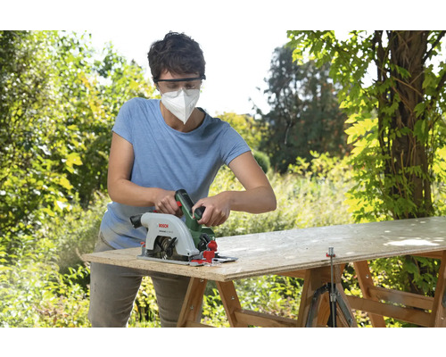Femme avec masque respiratoire coupant une planche en bois avec une scie circulaire à l'extérieur