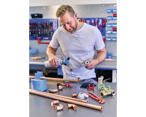 Homme soudant des tuyaux de cuivre avec un brûleur à gaz, entouré de raccords de tuyaux de cuivre et d''outils