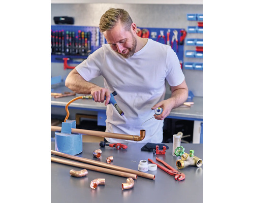 Un homme soude des tuyaux en cuivre avec un fer à souder dans un atelier. Divers outils et tuyaux sont posés sur la table.