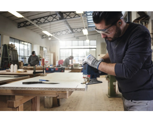 Un homme scie une planche de bois avec une scie sauteuse dans un atelier. Il porte des lunettes de protection et des gants.