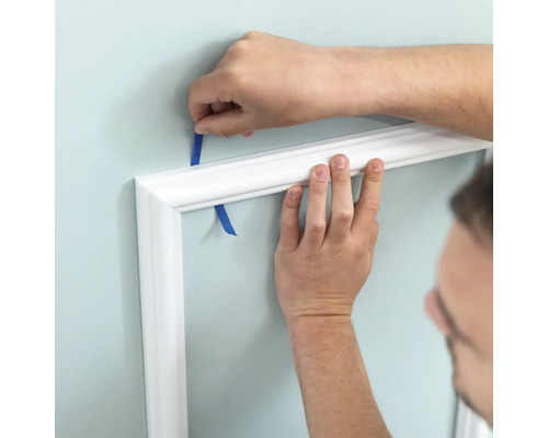 Person befestigt eine weiße Zierleiste mit Klebeband an der Wand.