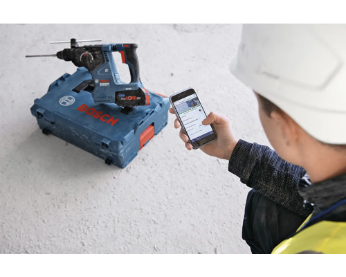 Perforateur à batterie Bosch sur une boîte à outils, un ouvrier avec un casque de protection regarde un téléphone portable