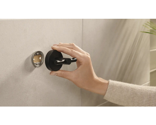 Eine Hand montiert einen schwarzen Doppelmantelhaken an einer Wandhalterung im Badezimmer.