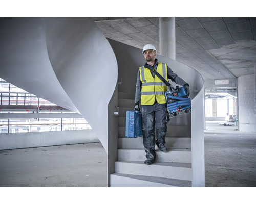 Ein Bauarbeiter mit Schutzhelm und Sicherheitsweste trägt eine Werkzeugkiste und einen Staubsauger die Treppe hinunter.