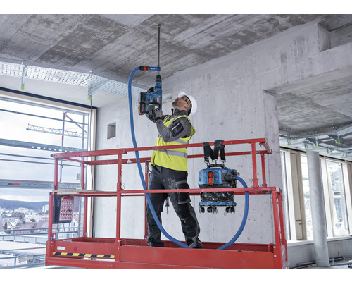 Ein Bauarbeiter bohrt mit einem Bohrhammer ein Loch in eine Betondecke. Ein Staubsauger ist mit dem Bohrhammer verbunden, um Staub zu entfernen.