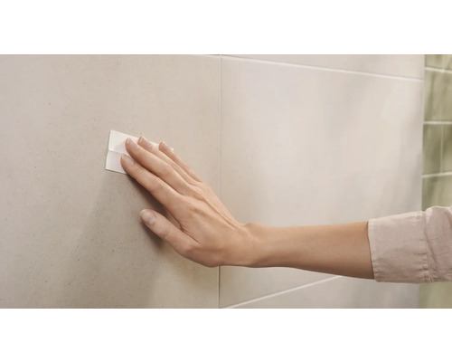 Eine Hand drückt ein Klebepad auf eine geflieste Wand
