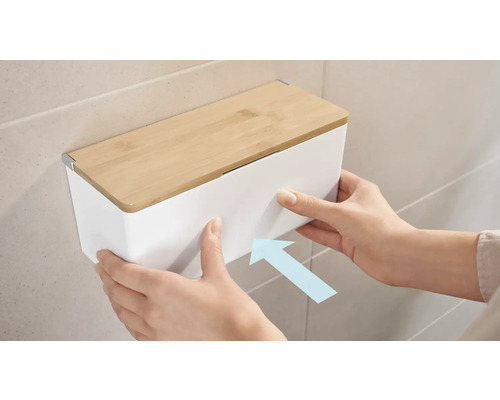 Eine Person montiert einen weißen Kosmetiktuchspender mit Bambusdeckel an der Wand.