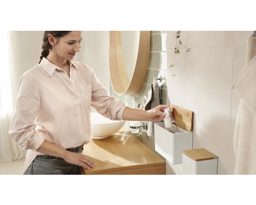 Salle de bains lumineuse avec lavabo, miroir et une femme qui sort une bouteille d''un rayonnage mural.