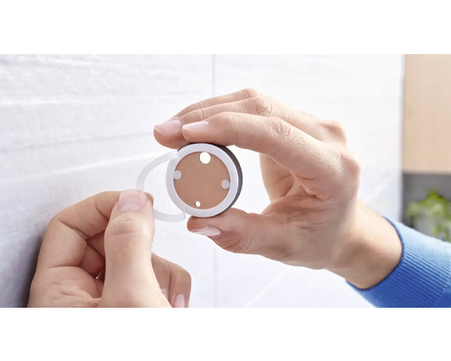 Eine Hand hält einen runden Wandhaken mit einem transparenten Befestigungselement zur Anbringung an einer Wand.