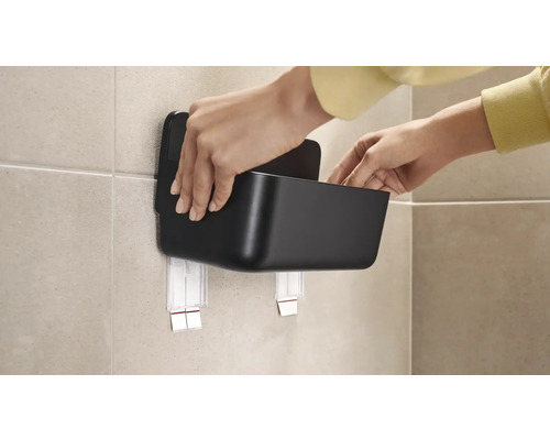 Person befestigt eine schwarze Aufbewahrungsbox mit Klebebefestigung an der Wand