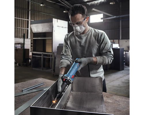 Mann mit Schutzbrille und Mundschutz verwendet einen Geradschleifer zur Metallbearbeitung in einer Werkstatt.