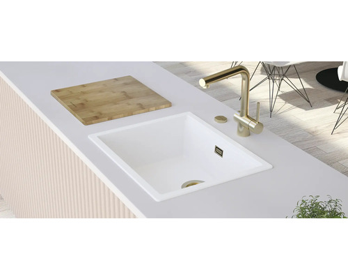 Scène de cuisine avec évier blanc, robinet doré et planche à découper en bois sur un plan de travail.