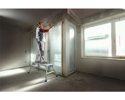 Artisan sur un marchepied lors de travaux de rénovation dans une pièce avec des fenêtres et des portes recouvertes.