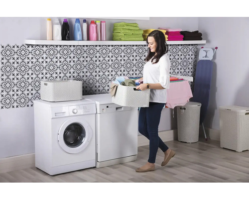 Buanderie lumineuse avec lave-linge, sèche-linge, panier à linge, coffre à linge, étagère et femme portant un panier à linge.