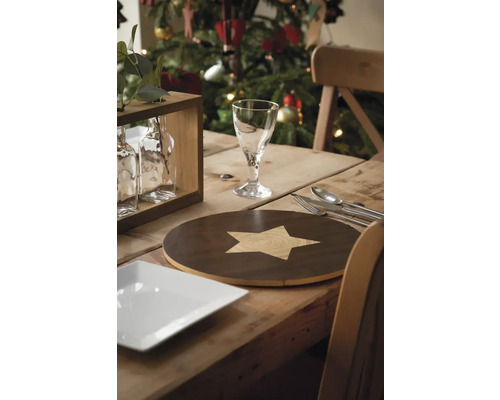 Table à manger décorée pour les fêtes avec un set de table à motif étoilé, un verre, des couverts et un sapin de Noël en arrière-plan.