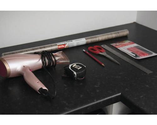 Sèche-cheveux, mètre ruban, ciseaux, crayon, règle et film pour meubles sur un plan de travail.