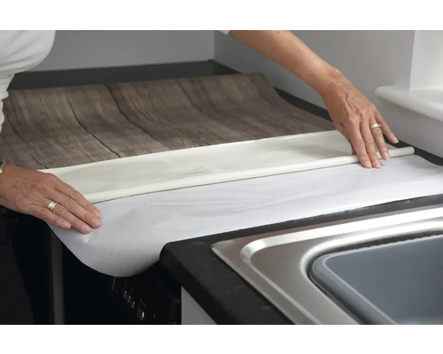 Une personne pose un film pour meubles imitation bois sur un plan de travail dans la cuisine.