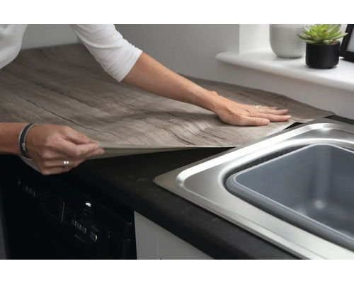 Scène de cuisine avec une personne appliquant un film décoratif autocollant imitation bois sur un plan de travail.