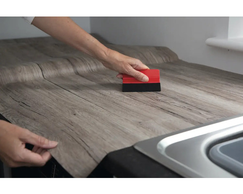 Une personne applique un film décoratif en bois sur un plan de travail à l''aide d''une raclette.