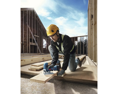 Un artisan portant un casque et des lunettes de protection est en train de scier une planche de bois avec une scie sauteuse sur un chantier.