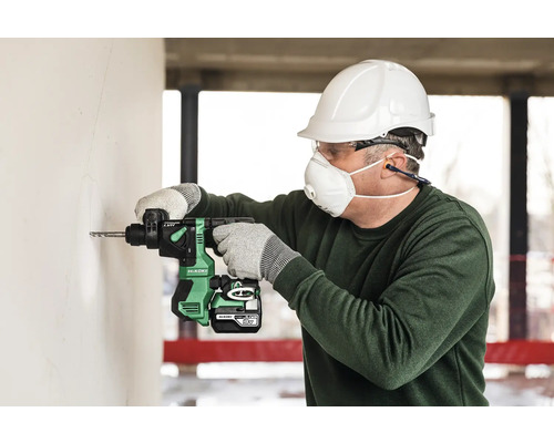 Ein Mann bohrt mit einem Akku-Bohrhammer ein Loch in eine Wand, wobei er Schutzkleidung wie Helm, Brille und Maske trägt.