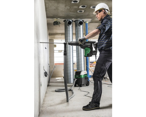 Un homme perce un trou dans un mur avec un marteau perforateur, tandis qu''un aspirateur industriel aspire la poussière.
