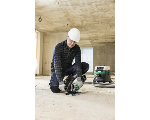 Un homme portant un casque et des lunettes de protection travaille avec une rainureuse à béton sur un sol en béton. Un aspirateur se trouve à l''arrière-plan.
