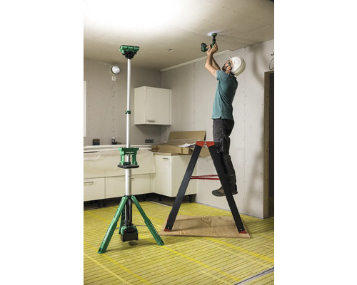 Innenaufnahme einer Baustelle mit einem Arbeiter auf einer Leiter, der eine Deckenlampe mit einem Akkubohrer installiert, im Vordergrund eine Baustellenleuchte