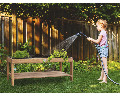 Ein Kind gießt Pflanzen in einem Hochbeet mit einem Gartenschlauch in einem Garten.