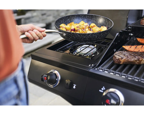 Une personne tient une poêle à frire contenant des pommes de terre sautées au-dessus d''un barbecue à gaz.
