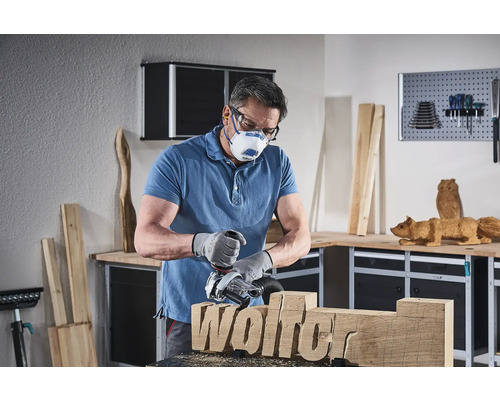 Un homme travaille avec une défonceuse sur une pièce de bois portant l''inscription Wolfer, tout en portant un masque de protection et des lunettes de protection dans un atelier.