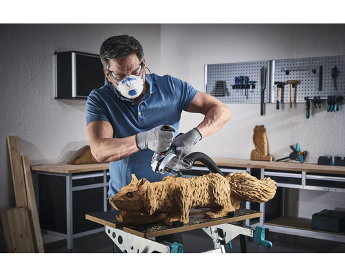 Un homme ponce une figurine d''écureuil en bois avec une ponceuse. Il porte un masque, des lunettes de protection et des gants.