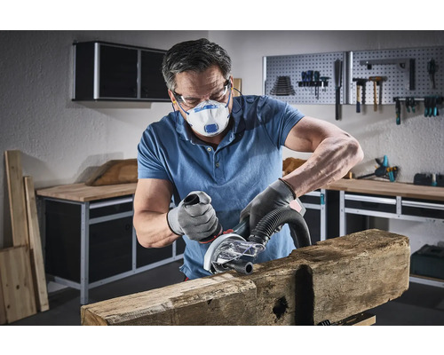 Un homme ponce une pièce de bois avec un outil électrique dans un atelier, portant un masque de protection et des gants.