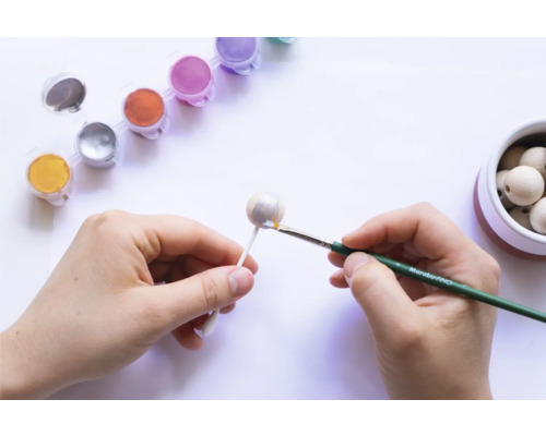 Une personne peint une boule en bois avec des couleurs et un pinceau. Des pots de peinture et d''autres boules en bois sont visibles sur l''image.