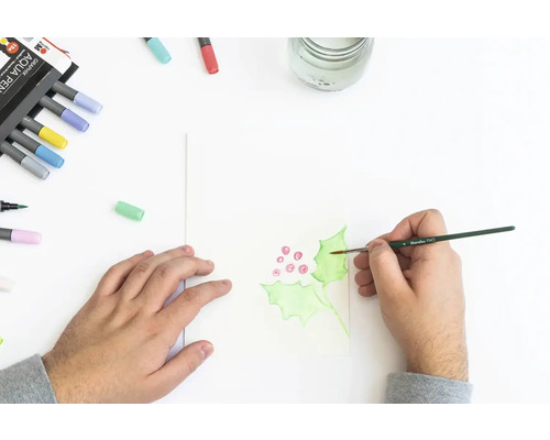 Une personne peint des feuilles de houx à l''aquarelle sur du papier. Des crayons aquarelle sont à côté.