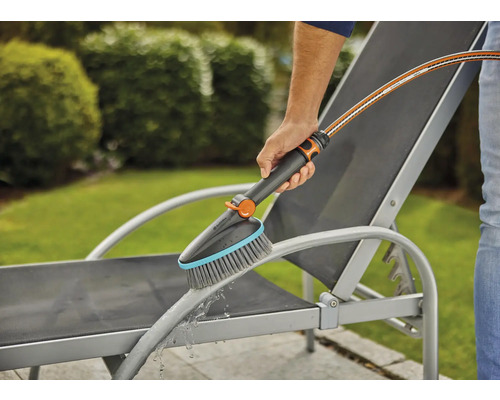 Une personne nettoie un transat de jardin avec une brosse et un tuyau d''arrosage.