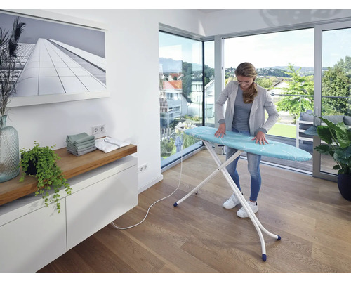 Frau bügelt Wäsche auf einem Bügeltisch in einem hellen Raum.