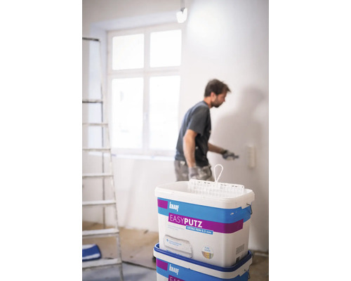 Un homme applique de l''enduit au rouleau sur un mur, seaux de Knauf Easyputz au premier plan