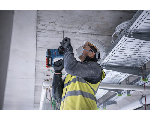 Ein Bauarbeiter bohrt mit einer Bohrmaschine in eine Betondecke, während er Schutzkleidung trägt.