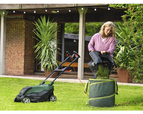Eine Frau leert Gras in einen Gartensack neben einem Rasenmäher auf einem Rasen.